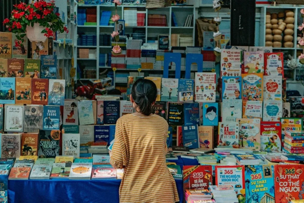 A woman browses a vibrant, crowded bookstall filled with colorful titles, symbolizing the challenge of standing out in a saturated book market.